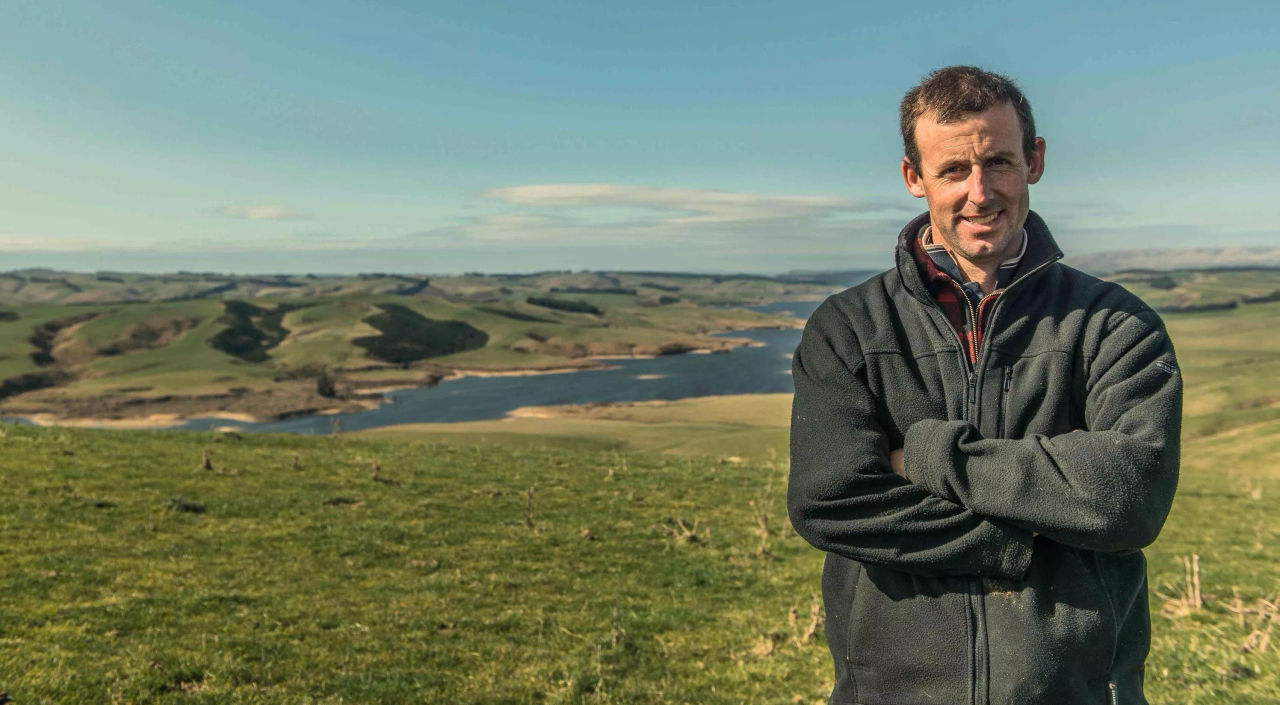 A smiling man stands with his arms folded in a paddock 